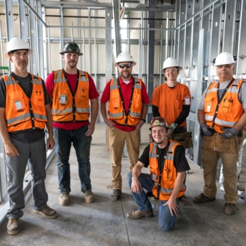 Six electricians in orange safety vests and hardhats stand in a building that is currently under construction.