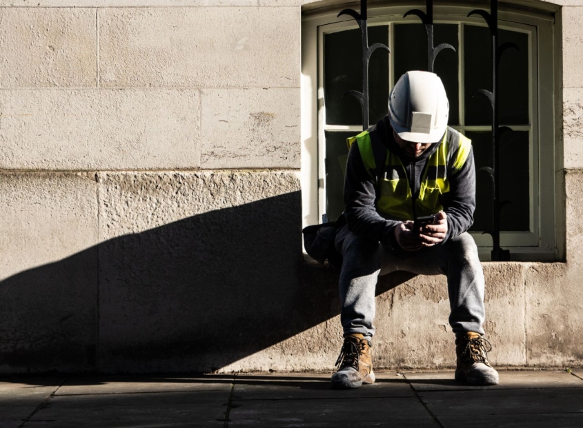 Electrical worker wearing a helmet and safety vest sits near a building while checking his smartphone.