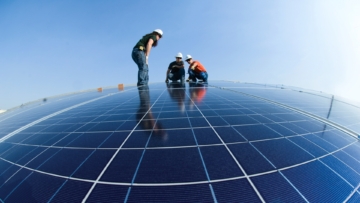Three electricians work to install solar panels on the roof of a commercial property.
