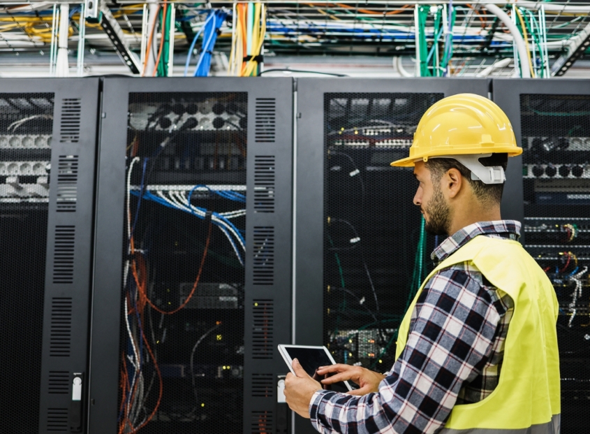 Commercial electrician in hard hat and safety vest checks the performance of a data center's servers.