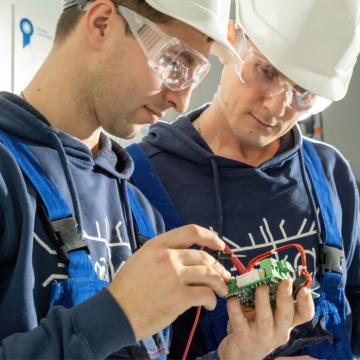 Two electricians wearing white helmets and blue sweatshirts examine an electrical box.