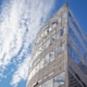 Modern office building with concrete exterior and large windows stands tall agains a background with a blue sky and clouds.