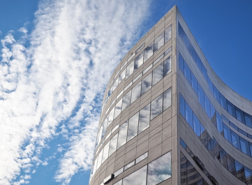 Modern office building with concrete exterior and large windows stands tall agains a background with a blue sky and clouds.