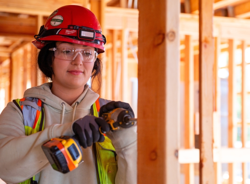 Woman construction worker wearing a red construction hat and using a drill on a house construction project.