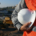 Construction worker in orange safety vest holding white hard hat outside a construction site.