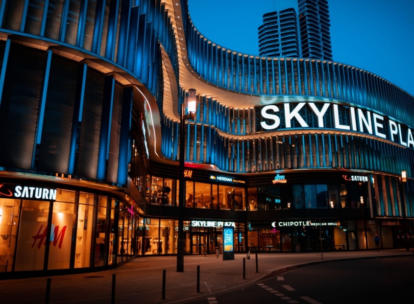 Nighttime exterior of Skyline Plaza with illuminated storefronts and wavy building facade.