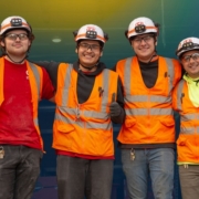 Four electrical workers smiling while wearing safety goggles, helmets, vest, and gloves.