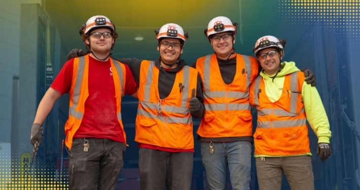 Four electrical workers smiling while wearing safety goggles, helmets, vest, and gloves.