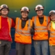 Four electrical workers smiling while wearing safety goggles, helmets, vest, and gloves.
