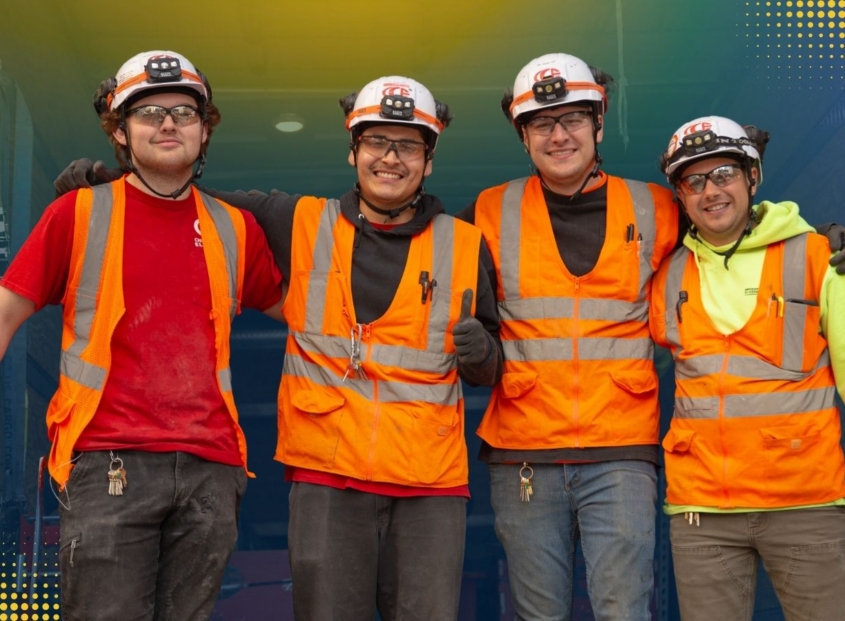 Four electrical workers smiling while wearing safety goggles, helmets, vest, and gloves.