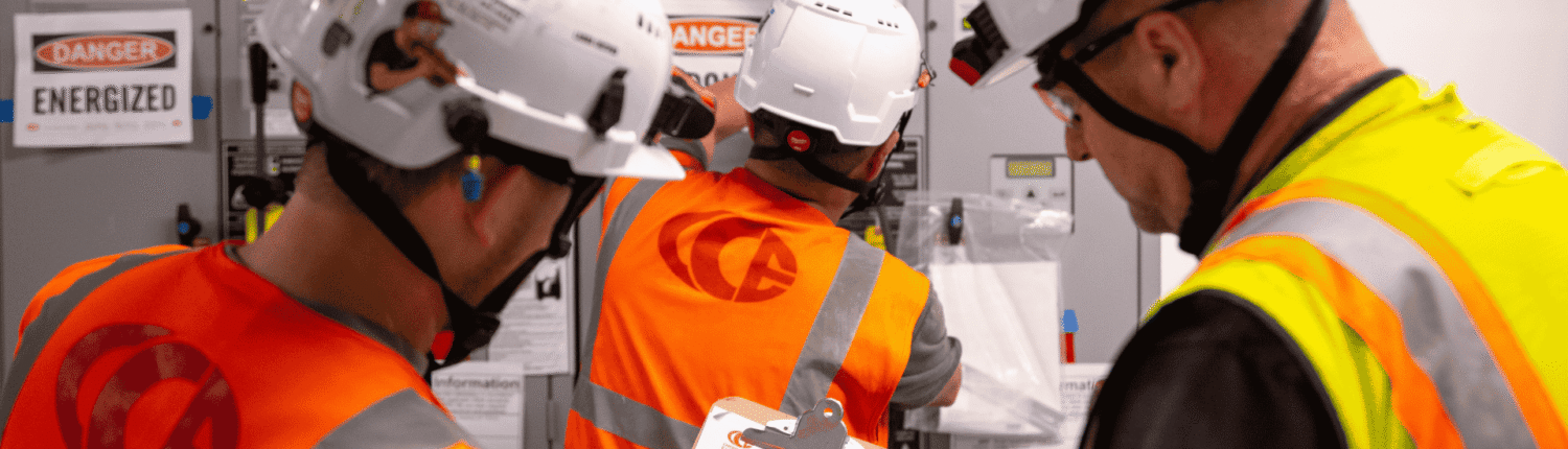 Three electricians in white hard hats and high-visibility vests reviewing a clipboard checklist in front of an energized electrical switchboard.