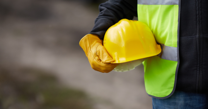 Close-up of a construction worker in a neon green safety vest and leather work gloves holding a yellow hard hat at a job site