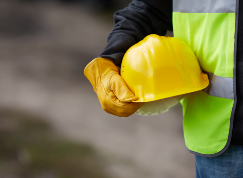Close-up of a construction worker in a neon green safety vest and leather work gloves holding a yellow hard hat at a job site