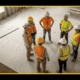 A group of construction workers in hard hats and high-visibility vests gathered in a circle on an active job site for a toolbox talk