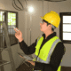 Electrician in a yellow hard hat and safety vest inspecting overhead wiring while holding a tablet inside a building under construction.
