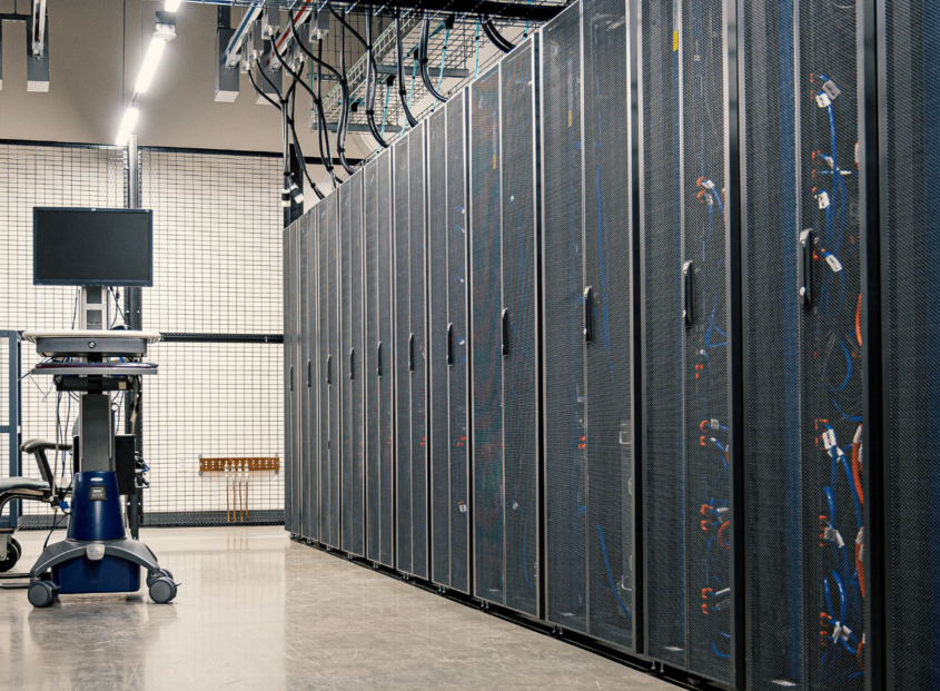 Interior of a data center with rows of server racks, cable management overhead, and a mobile workstation with a monitor in the aisle.
