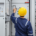 Electrician in a hard hat and safety coveralls operating switches on an industrial electrical control panel with rows of buttons and indicator lights.