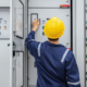 Electrician in a hard hat and safety coveralls operating switches on an industrial electrical control panel with rows of buttons and indicator lights.