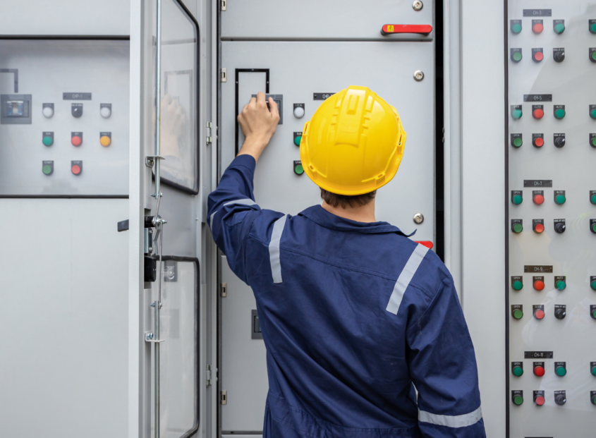 Electrician in a hard hat and safety coveralls operating switches on an industrial electrical control panel with rows of buttons and indicator lights.